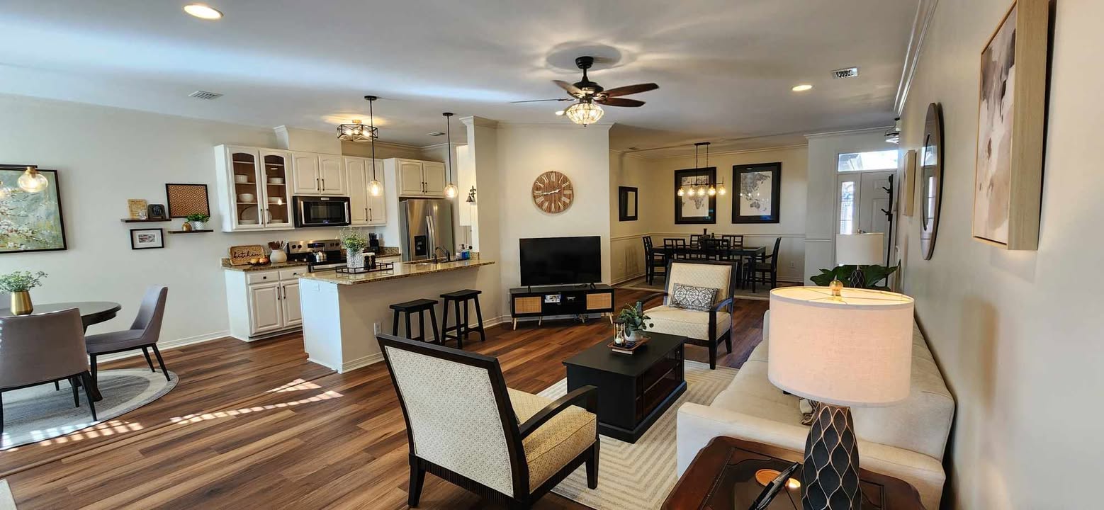 Warm mid-tone luxury vinyl plank flooring in an open-concept living room and kitchen with white cabinets, bar stools, dining table, and neutral furniture, shown clean and well-maintained with a smooth, matte finish and free of dust and debris.